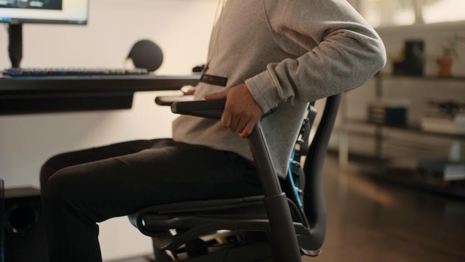 Ein Video von einer Person, die in einem schwarz-blauen Embody Gaming-Stuhl von Herman Miller sitzt und neben den Stuhl greift, um die Armlehnen zu verstellen.