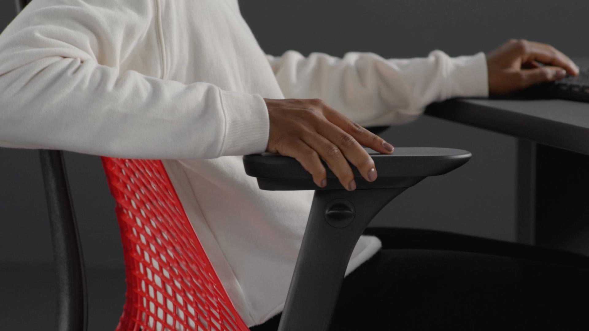 Ein Video von einer Person, die in einem rot-schwarzen Sayl-Gaming-Stuhl von Herman Miller sitzt und neben den Stuhl greift, um die Armlehnen zu verstellen.
