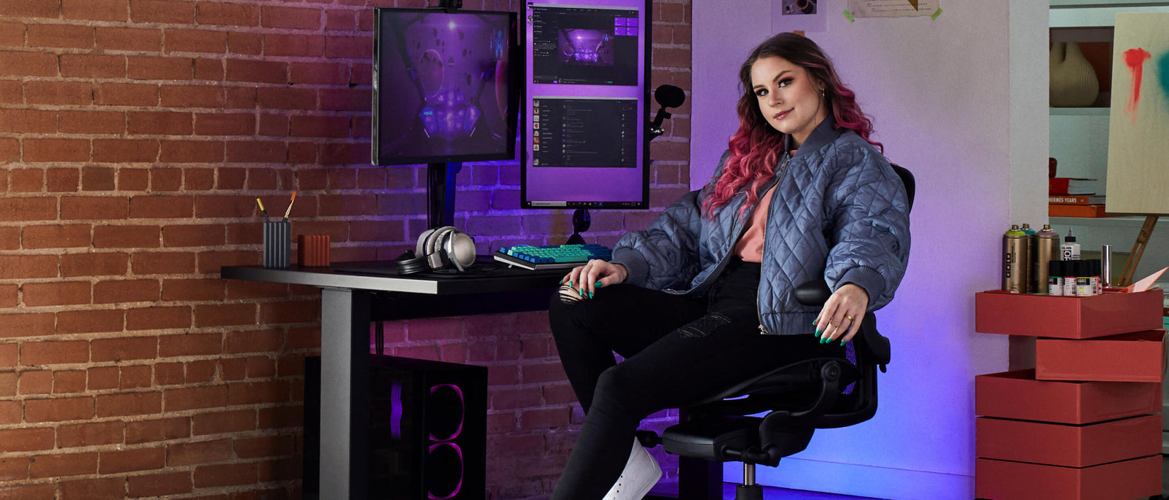 Foto der Gamerin Electra, die mit Blick zur Kamera auf einem schwarzen Aeron Gaming-Stuhl sitzt, vor einem schwarzen Nevi-Gaming-Tisch mit Ollin-Monitor