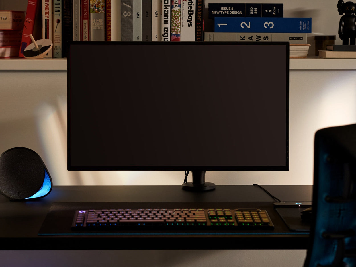 Vorderansicht eines schwarzen Ollin-Monitorarms, der einen Computerbildschirm hält und an einem schwarzen Nevi-Gaming-Schreibtisch von Herman Miller befestigt ist. Aufstellung in einem Spielzimmer oder Heimbüro, mit einem Bücherregal, Tastatur und Maus.