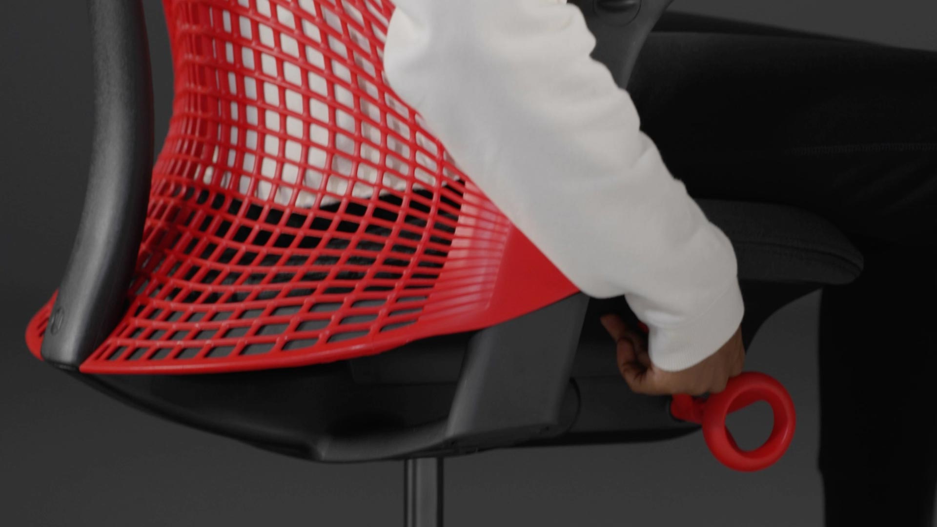 Ein Video von einer Person, die in einem rot-schwarzen Sayl-Gaming-Stuhl von Herman Miller sitzt und neben den Stuhl greift, um die Sitzhöhe einzustellen.