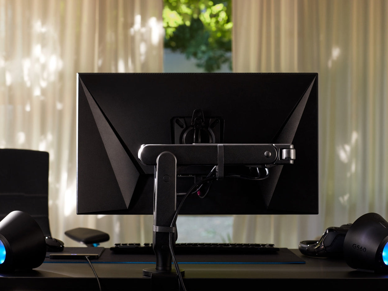 Rückansicht eines schwarzen Ollin-Monitorarms, der einen Computerbildschirm hält und an einem schwarzen Nevi-Gaming-Schreibtisch von Herman Miller befestigt ist. Set in einem Spielzimmer oder Heimbüro, mit Beleuchtung, Tastatur und Maus.