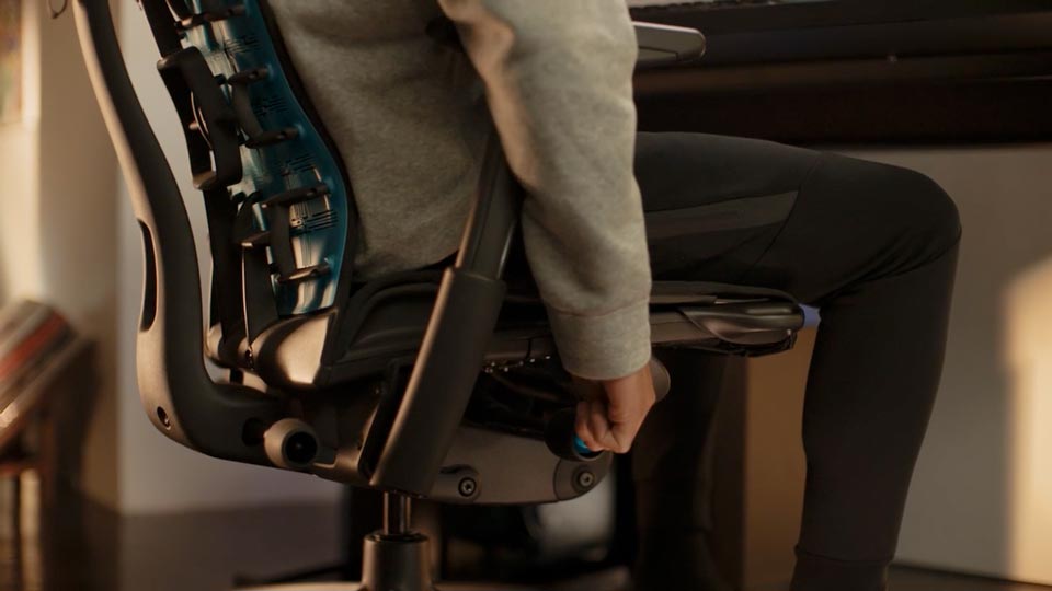 Ein Video von einer Person, die in einem schwarz-blauen Embody Gaming-Stuhl von Herman Miller sitzt und neben den Stuhl greift, um die Sitzhöhe einzustellen.