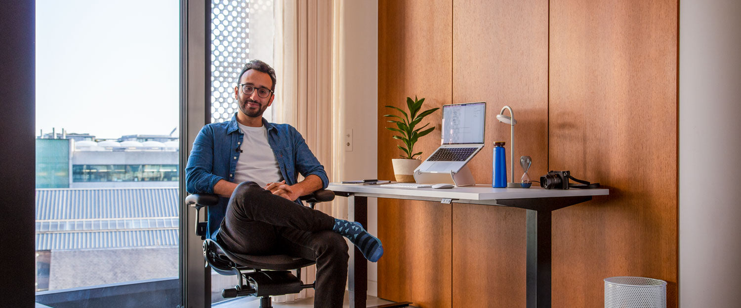 Foto von Ali Abdaal, Podcaster, sitzend auf einem Herman Miller Aeron Bürostuhl in Graphit vor einem Nevi Steh-Sitz-Schreibtisch mit schwarzen Beinen und weißer Platte, mit einem Laptop in einem Oripura Laptopständer