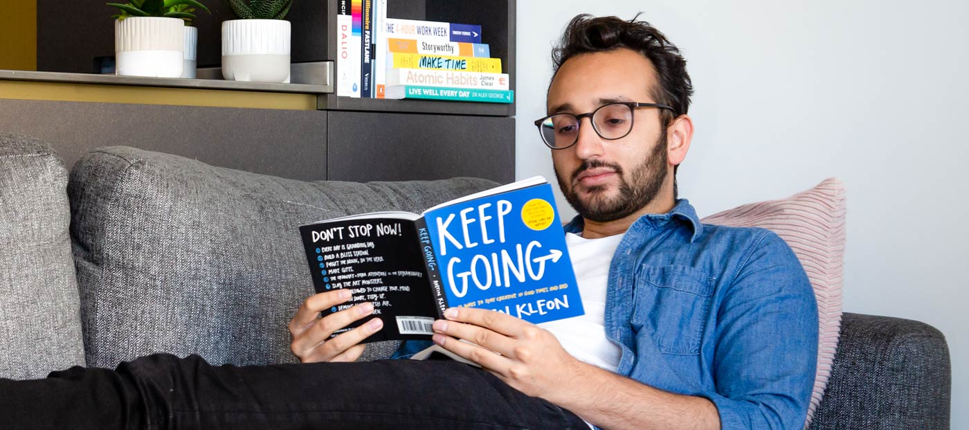 Foto von Ali Abdaal auf dem Sofa liegend, ein Buch mit dem Titel „Keep Going” lesend
