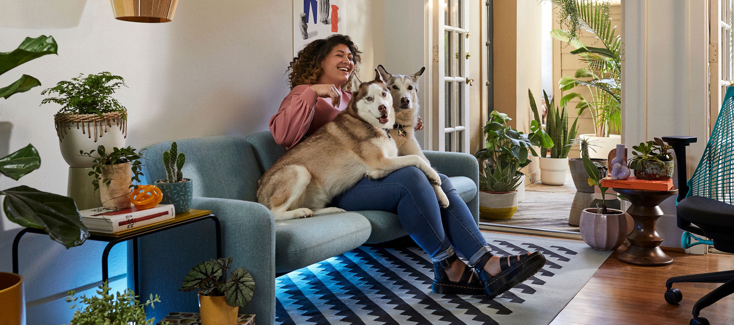 Friskk sitzt auf einem grünen Wilkes-Sofa mit ihren beiden Hunden, neben ihr stehen ein schwarzer und ein ozeanblauer Sayl-Gaming-Sessel