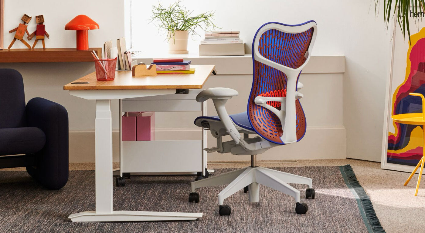 Ein blauer, orangefarbener und weißer Mirra 2 Bürostuhl von Herman Miller, der neben einem Jarvis Stehpult steht. In einer Home-Office-Umgebung mit einem blauen Wilkes-Sofa, einem blauen Maharam-Teppich, einem gelben DLM-Beistelltisch und einer roten HAY Pao-Tischleuchte.