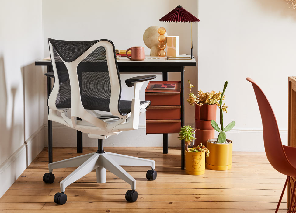 Ein blau-weißer Cosm-Bürostuhl von Herman Miller vor einem schwarz-terracottafarbenen Mode-Schreibtisch von Herman Miller. In einem Heimbüro mit Holzboden und weißen Wänden, ausgestattet mit einer Matin-Tischleuchte und Zimmerpflanzen.