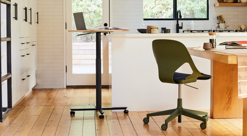 Ein schwarzer und eichenfarbener Passport-Tisch von Herman Miller, der neben einer weißen Kücheninsel steht, mit einem Laptop und einem Buch. Das Bild zeigt auch einen grün-schwarzen Zeph-Stuhl von Herman Miller, der an einem Esstisch aus Holz steht.