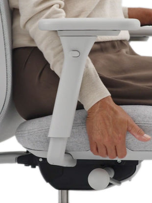 Seitliche Nahaufnahme einer Person, die auf einem grauen Lino-Bürostuhl von Herman Miller sitzt. Sie ist mit einem beigen Pullover und einer braunen Hose bekleidet und nutzt die Funktion zur Einstellung der Sitztiefe.