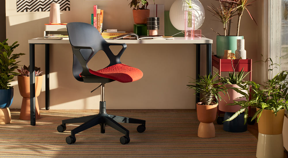 Ein schwarzer Zeph-Bürostuhl mit einem roten Sitzpolster steht vor einem Herman Miller OE1-Schreibtisch. In einer Home-Office-Umgebung, mit einem roten OE1-Trolley und einer Auswahl an Zimmerpflanzen in bunten Töpfen.