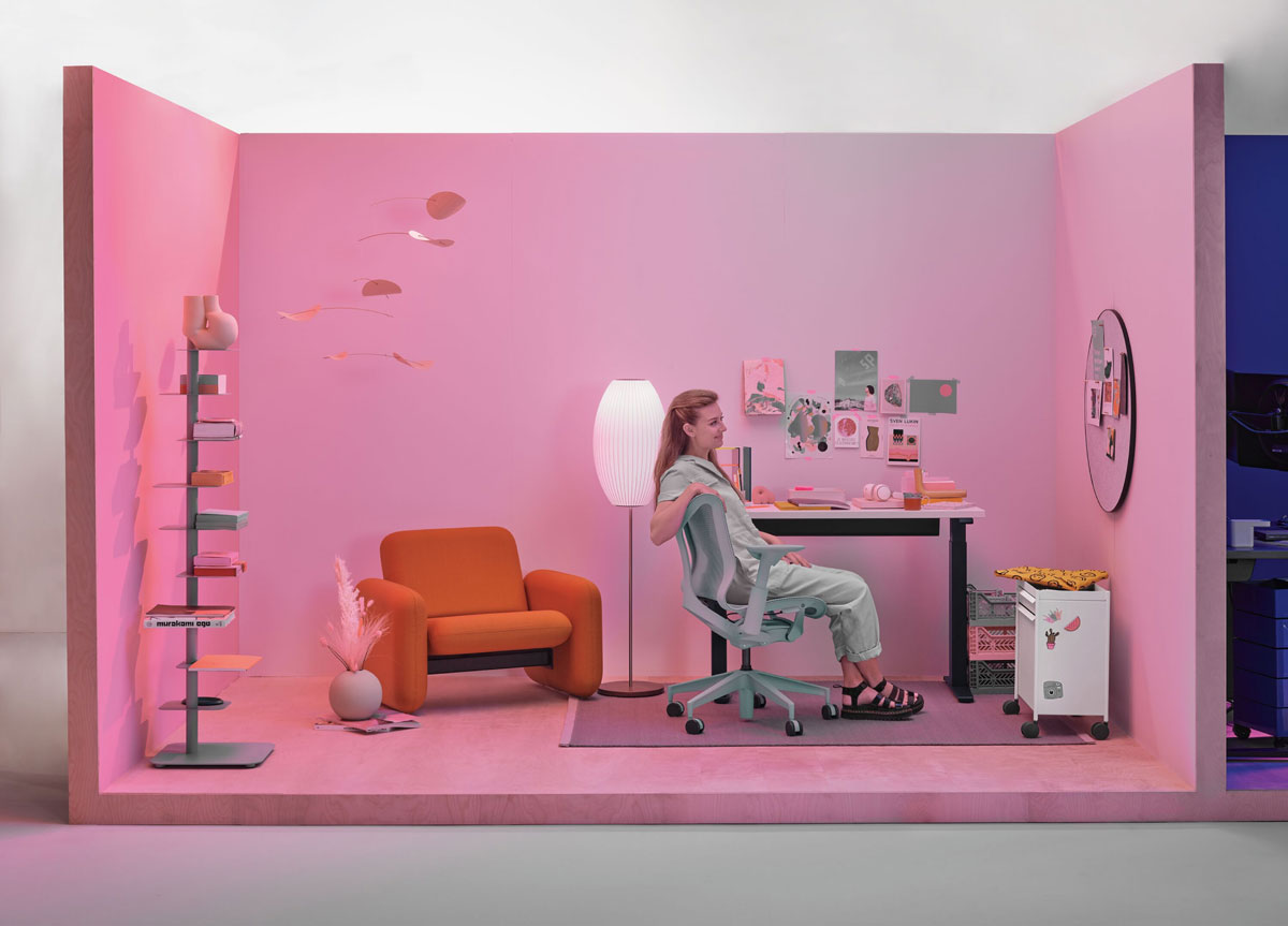 Homeoffice mit Frau, die in einem Cosm-Bürostuhl mit niedriger Rückenlehne in Gletschergrau sitzt, mit kompaktem Steh-Sitz-Schreibtisch und Wilkes-Armlehnstuhl im Hintergrund