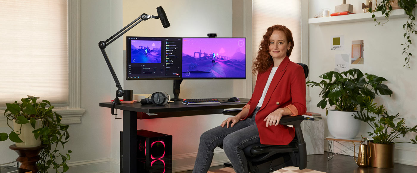 Foto von Dr. Lindsey Migliore, die in einem Embody-Gaming-Stuhl sitzt und in die Kamera blickt, mit einem schwarzen Nevi-Gaming-Schreibtisch und Monitorarmen von Ollin