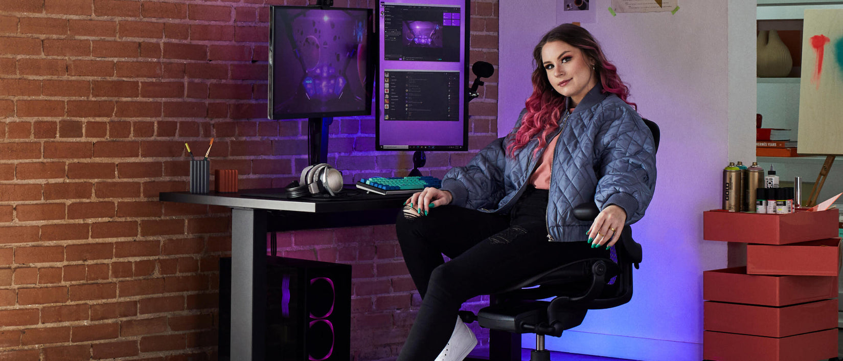 Foto der Gamerin Electra, die mit Blick zur Kamera auf einem schwarzen Aeron Gaming-Stuhl sitzt, vor einem schwarzen Nevi-Gaming-Tisch mit Ollin-Monitor