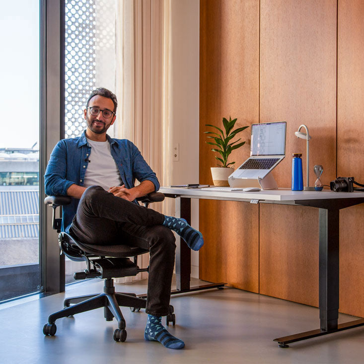 Foto von Ali Abdaal, Podcaster, auf einem Herman Miller Aeron-Bürostuhl in Graphit vor einem Nevi-Sitz-Steh-Schreibtisch mit schwarzen Beinen und weißer Platte mit einem Laptop in einem Oripura-Laptopständer
