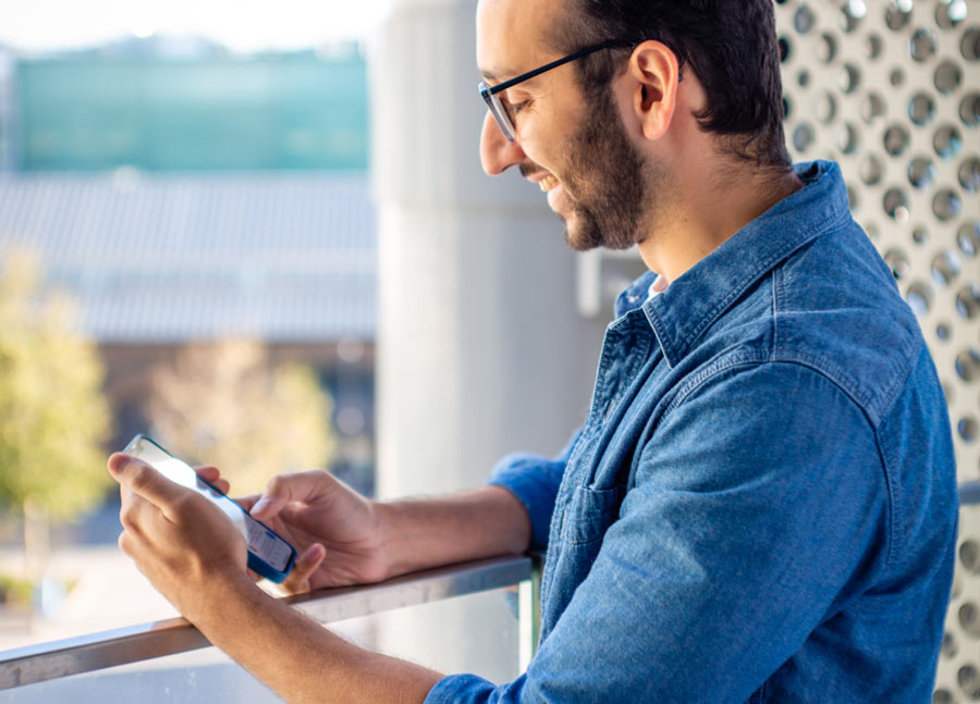 Foto von Ali Abdaal am Fenster in seinem Zuhause, auf sein Handy schauend