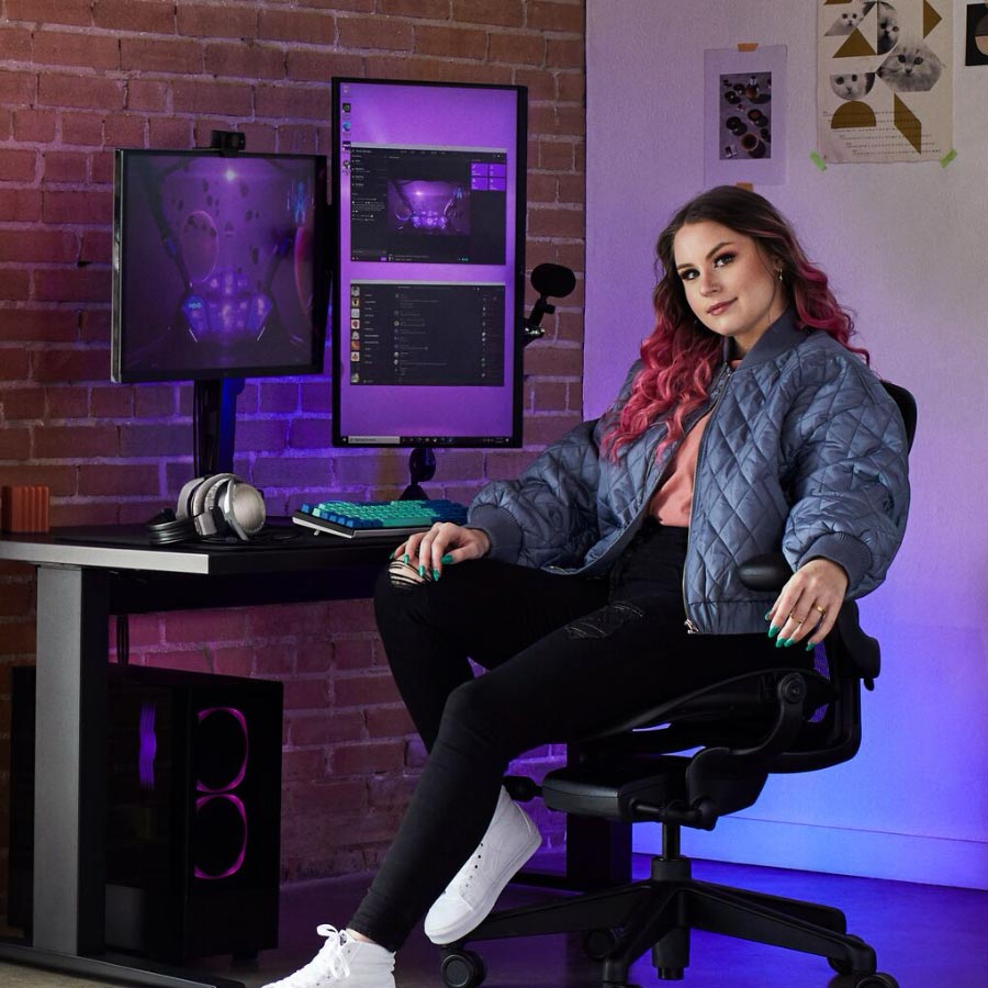 Star-Gamer und Coach Electra saß an einem Gaming-Set, das einen Herman Miller Aeron Gaming-Stuhl mit einem Nevi-Sitz-Steh-Schreibtisch in Schwarz mit zwei Monitoren auf Ollin-Monitorarmen enthielt