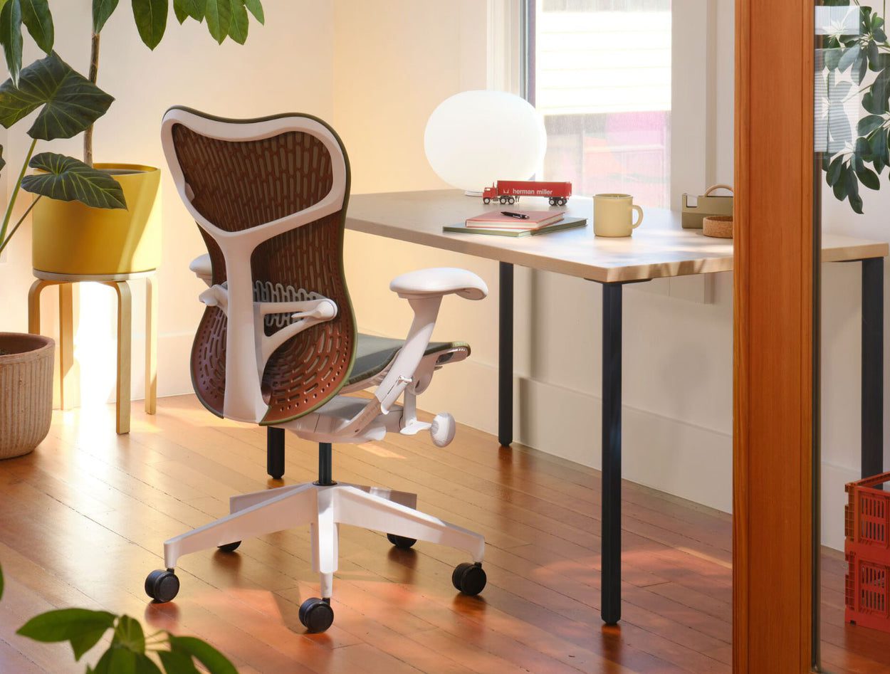 Ein olivgrüner und weißer Mirra 2-Bürostuhl mit kastanienbrauner Rückenlehne, der vor einem Herman Miller OE1-Schreibtisch steht. In einer Home-Office-Umgebung mit Massivholzböden, weißen Wänden und warmer, gemütlicher Beleuchtung.