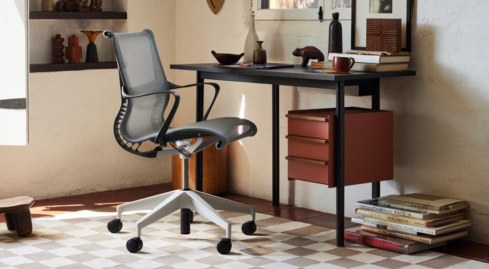 Ein schwarzer Mode-Schreibtisch von Herman Miller mit Ablageflächen in Terrakotta und Griffen aus Nussbaumholz steht vor einem grauen Setu-Bürostuhl von Herman Miller. In einem gut eingerichteten Heimbüro mit Regalen, Büchern und dekorativen Wohnaccessoires.