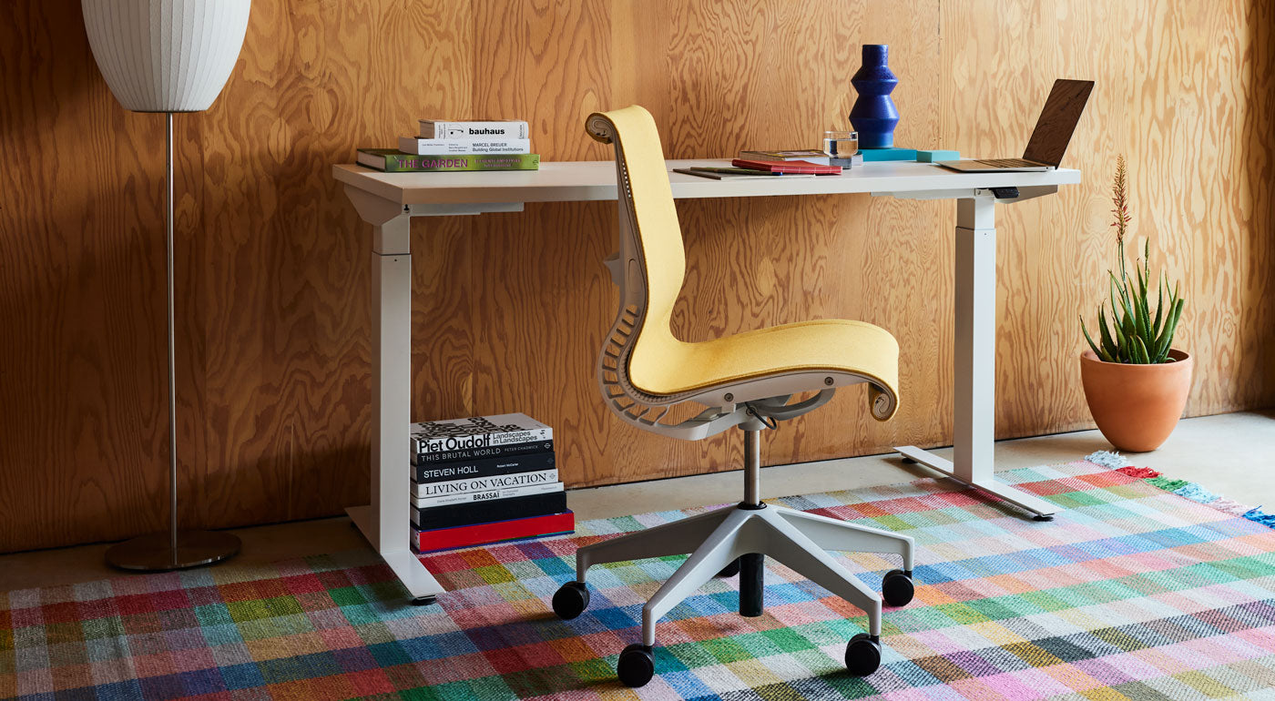 Ein gelber Setu-Bürostuhl von Herman Miller, bezogen mit dem Stoff Mimosa Meld, steht vor einem weißen Nevi-Stehpult auf einem bunten Karoteppich.