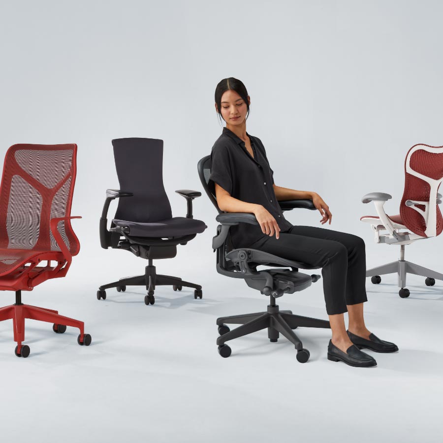 Aufstellung von vier Herman Miller-Bürostühlen, darunter Cosm Mid Back in Canyon Rot, Embody in Schwarz, Aeron in Graphit, auf dem eine Dame sitzt, und schließlich Mirra 2 mit weißem Gestell und rotem Sitz und roter