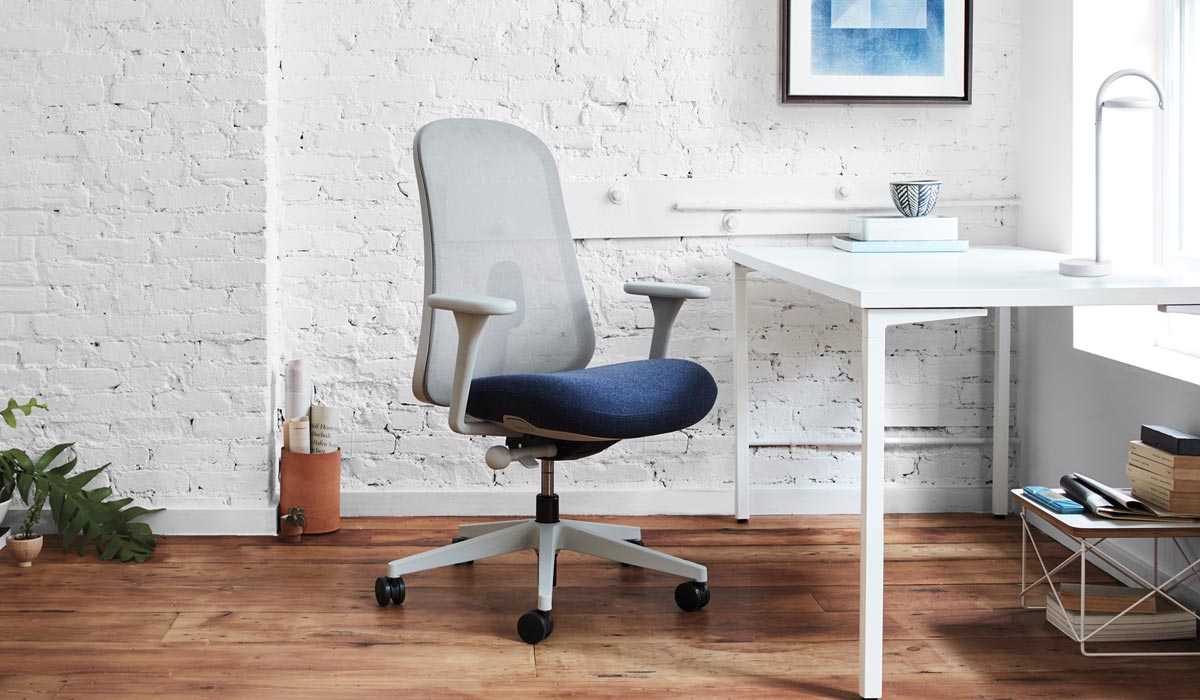 Ein grauer und blauer Lino-Bürostuhl von Herman Miller, der vor einem weißen Schreibtisch steht. Die Inneneinrichtung besteht aus einer weißen Backsteinwand, einem Holzfußboden und einer weißen Marselis-Tischleuchte auf dem Schreibtisch.