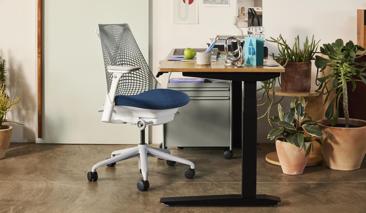 Ein weiß-blauer Sayl-Bürostuhl von Herman Miller neben einem Jarvis-Stehpult mit schwarzen Beinen und einer Arbeitsplatte aus Bambus. In einer Home-Office-Umgebung, mit einem blauen OE1-Trolley und einer Auswahl von Zimmerpflanzen in Terrakotta-Töpfen.