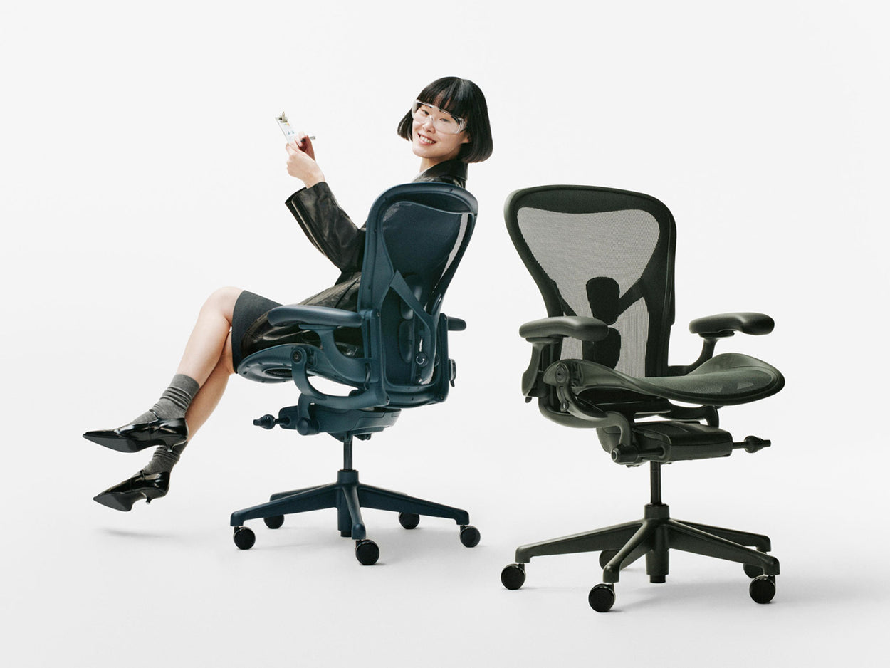 In einem weißen Studio saß eine Frau auf einem ergonomischen Bürostuhl vom Typ Herman Miller Aeron in der Farbe „Nighfall Blue“ neben einem weiteren Aeron in der Farbe „Jasper Green“.