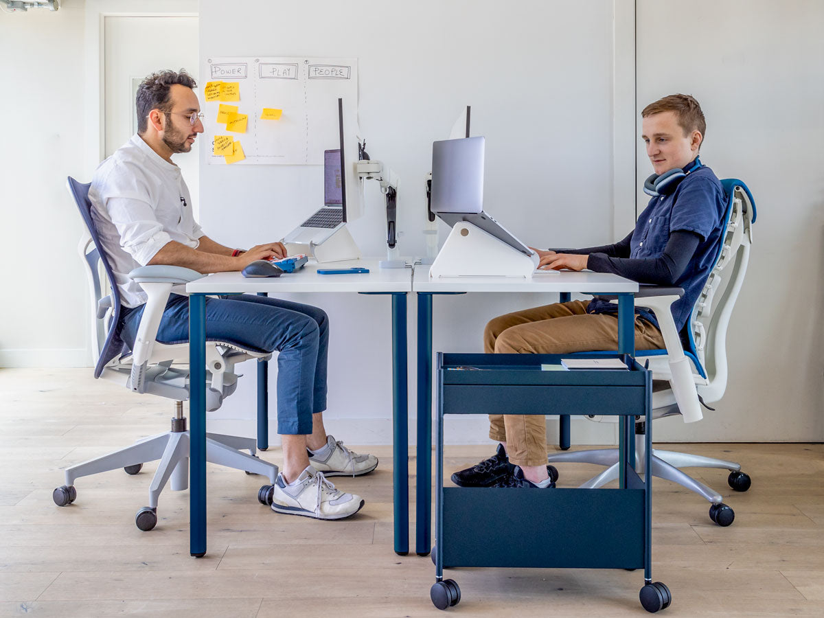Foto von 2 Männern, die sich am Schreibtisch gegenübersitzen, das zeigt, dass die Ellbogen beim korrekten Sitzen auf gleicher Höhe mit der Tischplatte sind. Verfügt über einen OE1-Schreibtisch mit blauen Beinen und weißer Platte zusammen mit Mirra 2 in Mineralweiß und Blau und Embody in Weiß und Blau