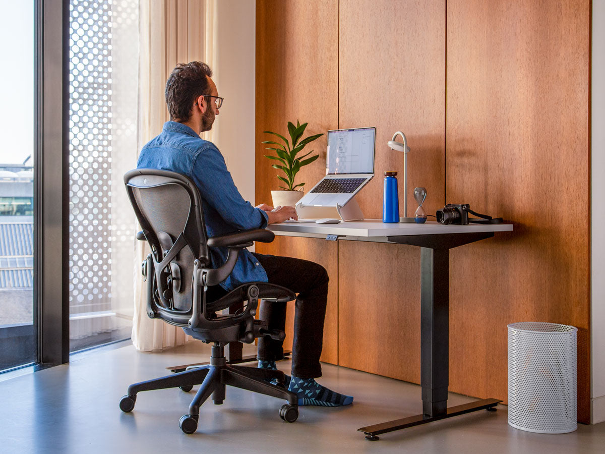 Foto eines Mannes, der in einem Aeron-Bürostuhl in Graphit sitzt, der korrekt zeigt, dass die Füße auf dem Boden stehen sollten, wenn man richtig sitzt