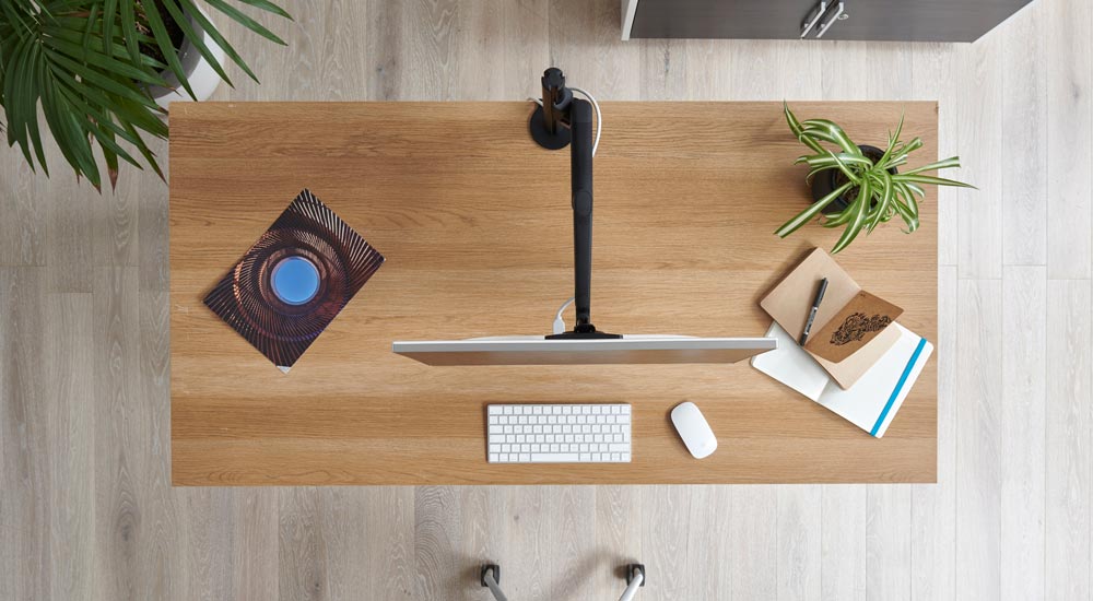 Draufsicht auf einen schwarzen Lima-Monitorarm von CBS, der an einem Holzschreibtisch befestigt ist und die Bewegung von Monitorarm und Bildschirm zeigt.