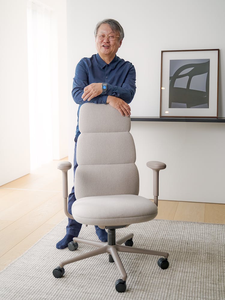 Der Designer Naoto Fukasawa lehnt sich an einen Asari-Bürostuhl von Herman Miller. Die Umgebung besteht aus einem sauberen Boden und einer Wand im Hintergrund.