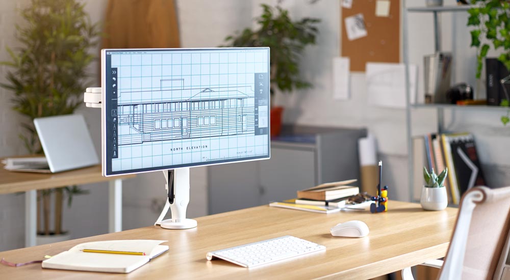 Ein weißer Ollin-Monitorarm von CBS, montiert auf einem Holzschreibtisch mit einem Computerbildschirm, in einer Büroumgebung.