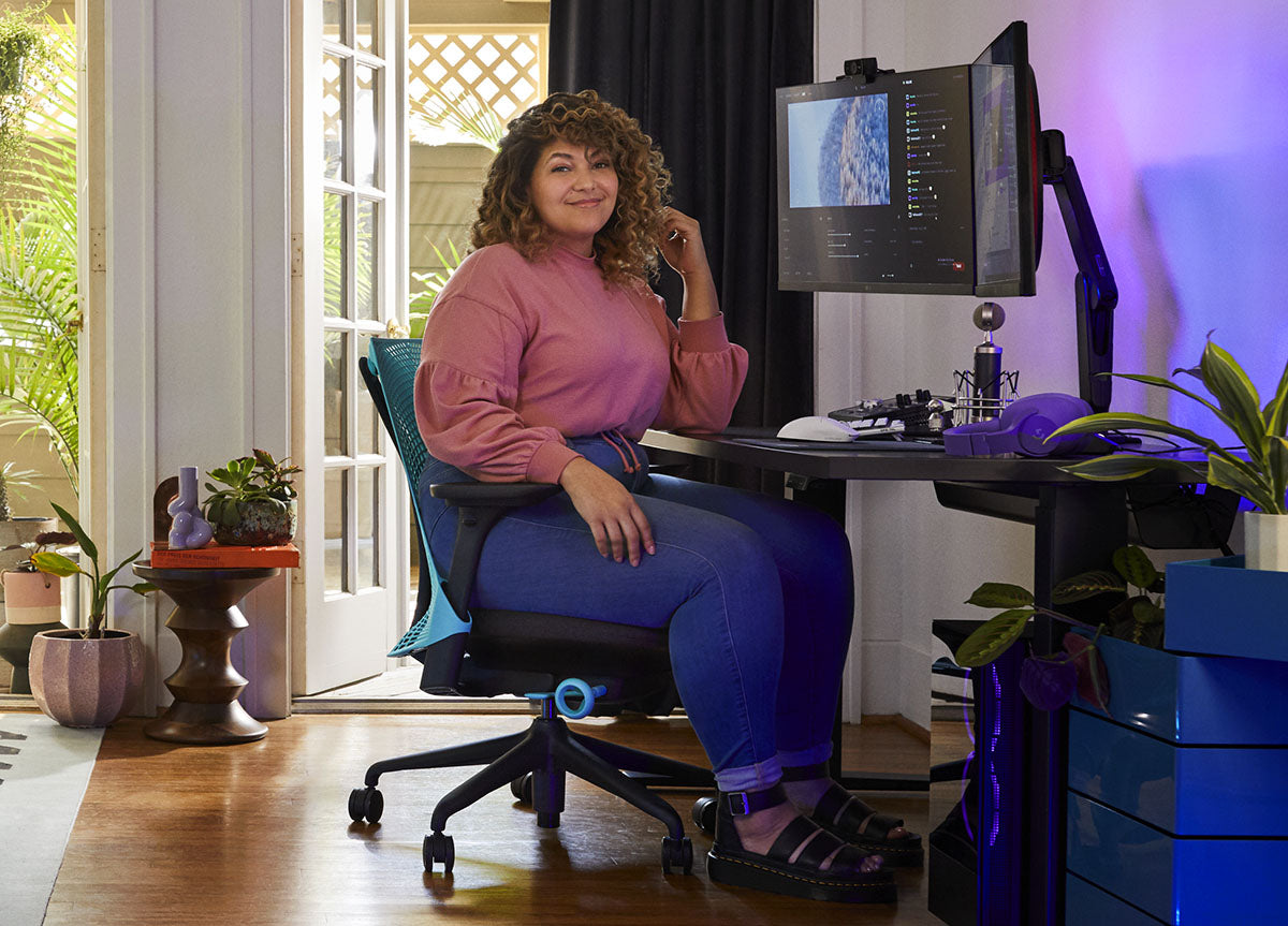 Eine Frau sitzt in einem blau-schwarzen Sayl-Gaming-Stuhl von Herman Miller hinter einem schwarzen Nevi-Schreibtisch mit einem schwarzen Ollin-Monitorarm und -Bildschirm. In einem Innenraum mit Holzböden, weißen Wänden und Zimmerpflanzen.