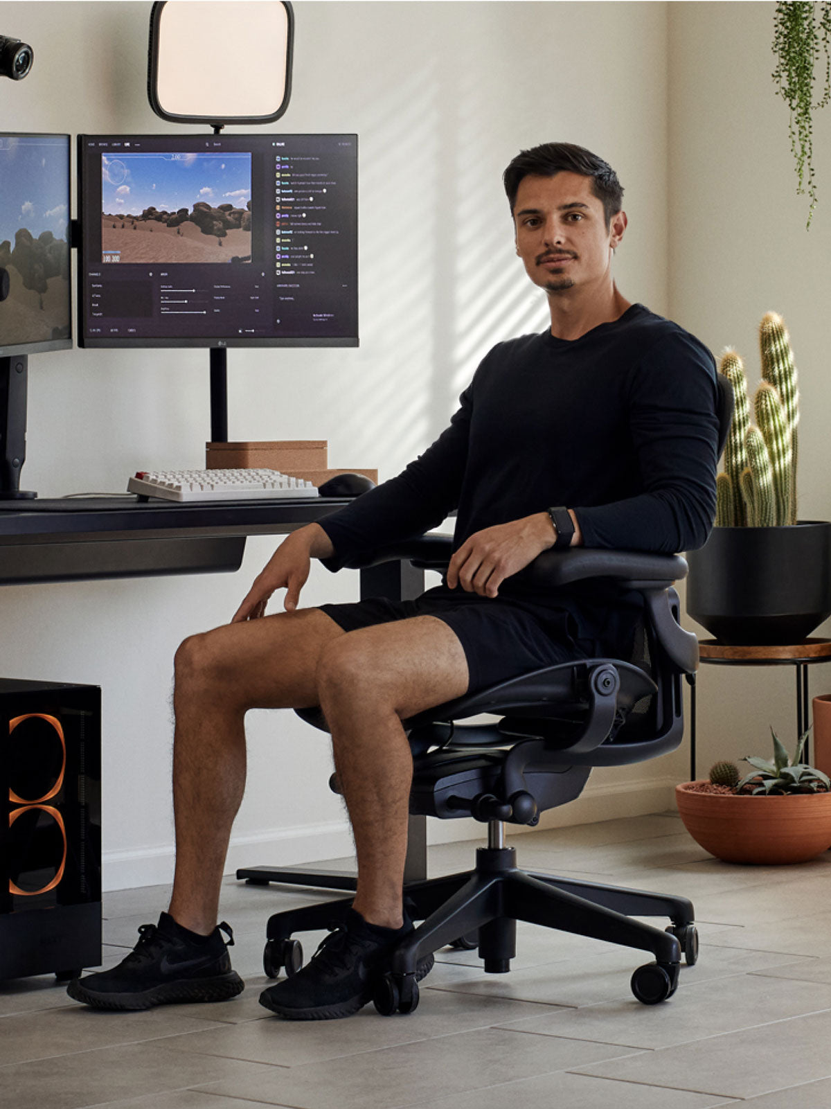 Ein Mann sitzt in einem schwarzen Aeron-Gaming-Stuhl von Herman Miller hinter einem schwarzen Nevi-Schreibtisch mit einem schwarzen Ollin-Monitorarm und Bildschirm. In einem Innenraum mit gefliesten Böden, weißen Wänden und Zimmerpflanzen in Terrakotta-Töpfen.