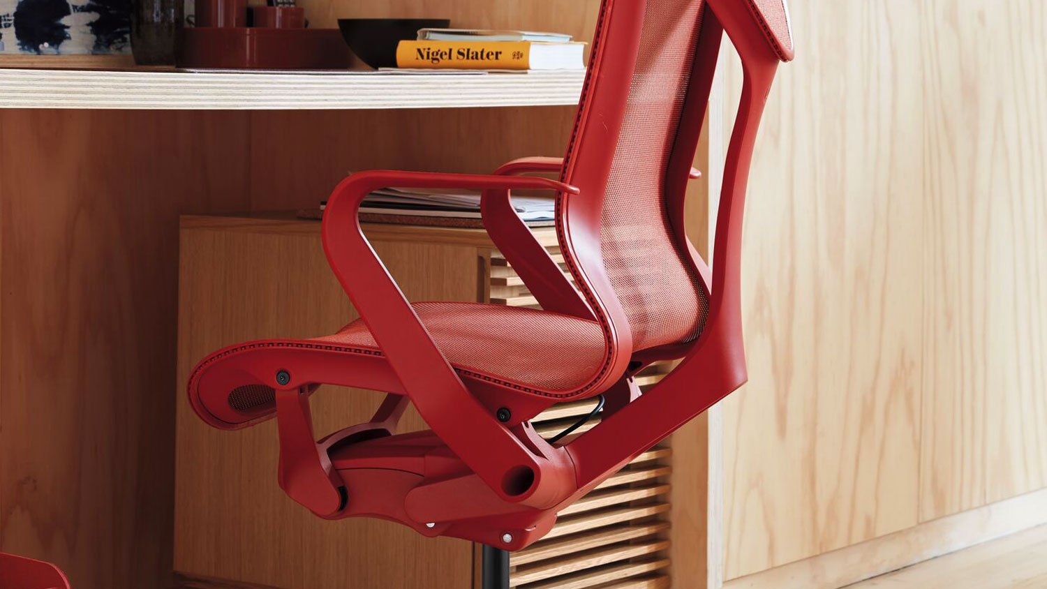 Ein roter Cosm-Bürostuhl von Herman Miller, der unter einem Schreibtisch steht. Er steht in einem Innenraum mit Hartholzböden und -wänden, wobei der Schwerpunkt auf der leuchtenden Farbe des Stuhls inmitten der umgebenden Möbel liegt.