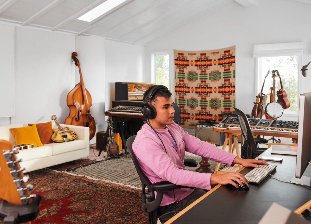 Rostam, Musiker, Produzent und Komponist, sitzt mit Kopfhörern am Computer auf einem Aeron-Bürostuhl in Graphit, im Hintergrund sind Musikinstrumente zu sehen.